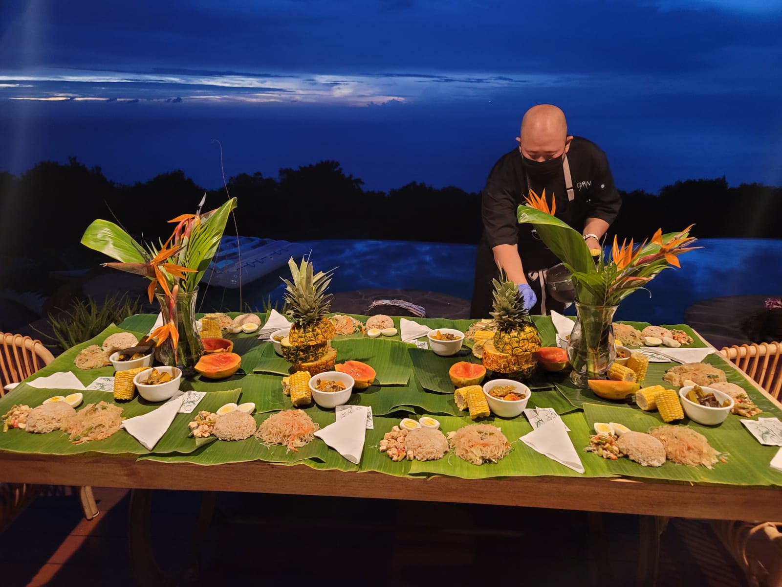 Outdoor Kamayan-style table at night beside a pool with Chef Dayne serving